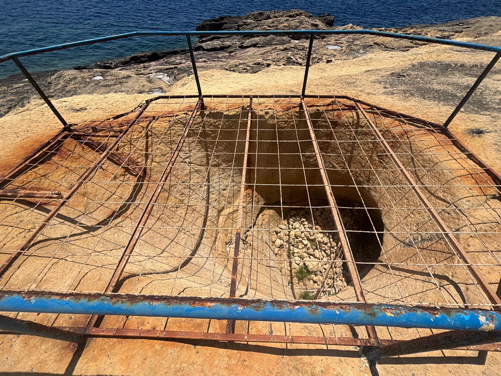 High angle view looking straight down through a rusted iron safety grid into the circular mouth of the rock mortar