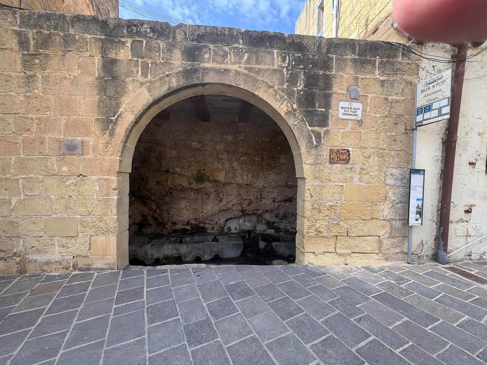 Front view of L-Ghajn ta Bendu wash house arch in Fontana Gozo with stone paving and a bus stop sign to the right