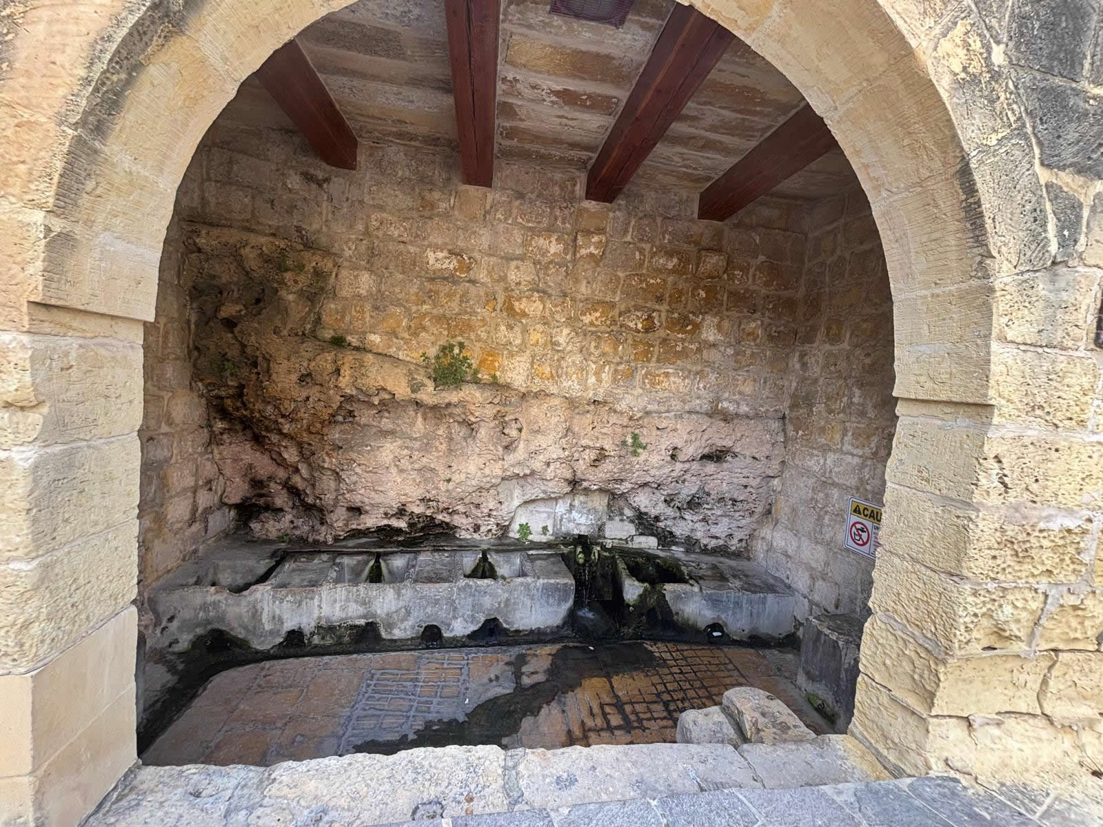 Interior view of Ghajn ta Bendu showcasing restored timber ceiling beams and stone basins built against the exposed bedrock