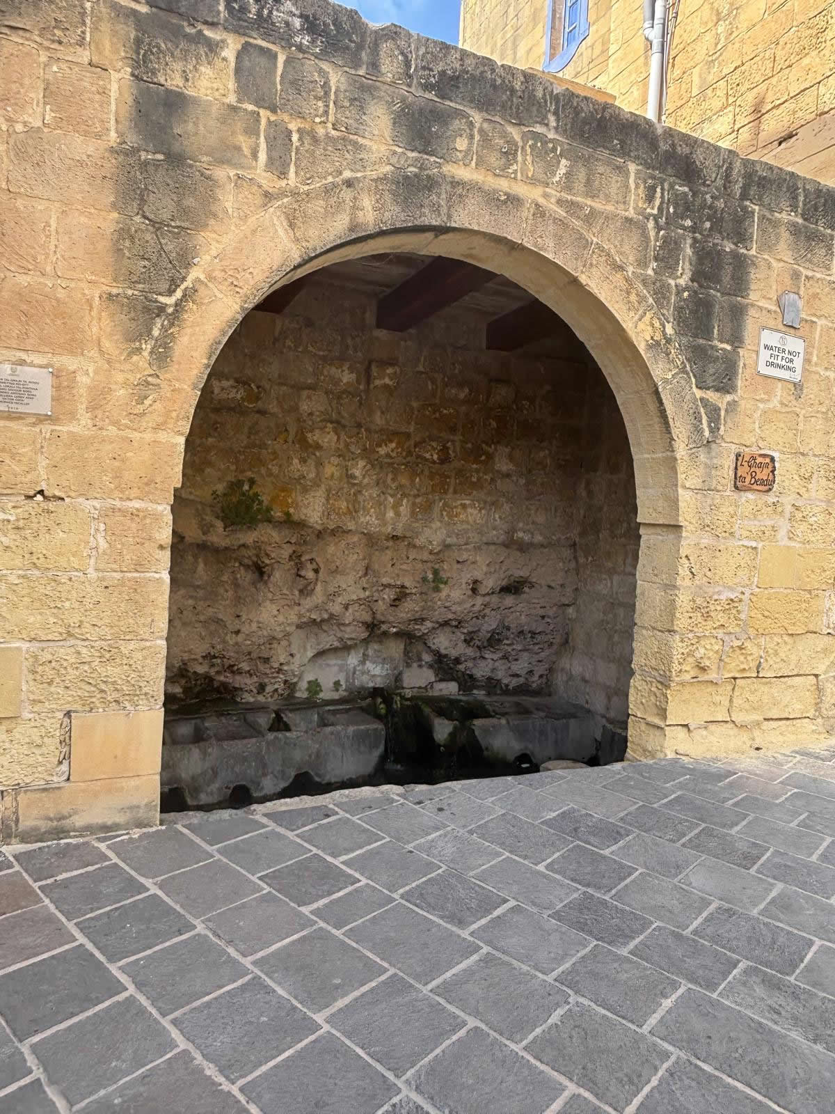 Angled exterior street view of the limestone arch of L-Ghajn ta Bendu in Fontana Gozo