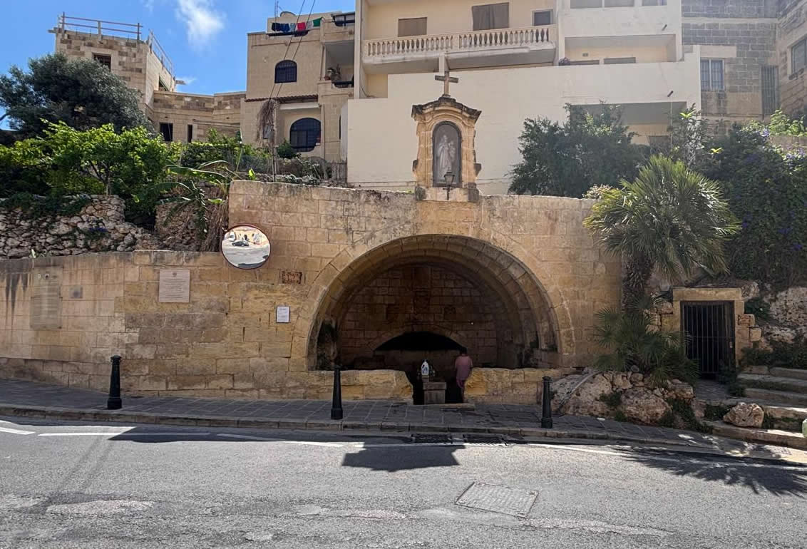 Street view of the large stone archway of L-Ghajn il-Kbira wash house beneath a statue niche and modern apartments in Fontana Gozo