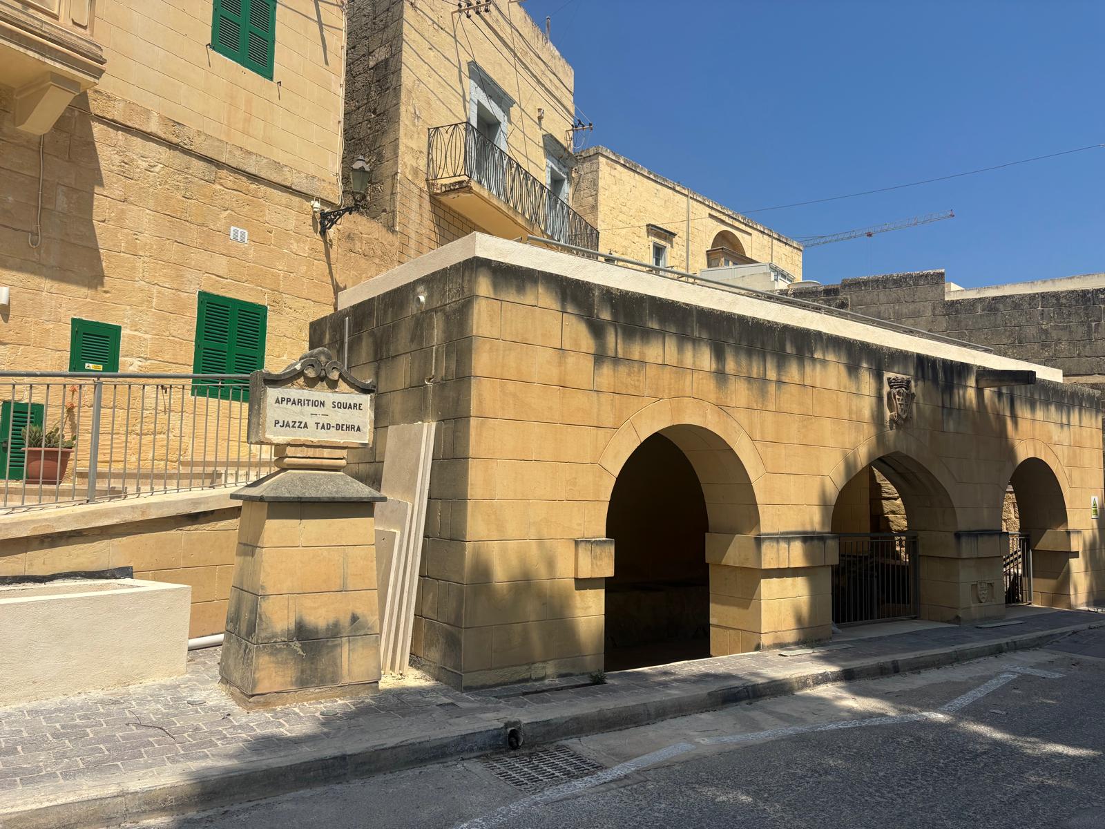 The historic stone arched wash house structure at Pjazza tad-Dehra in Għajnsielem, with traditional limestone buildings behind it