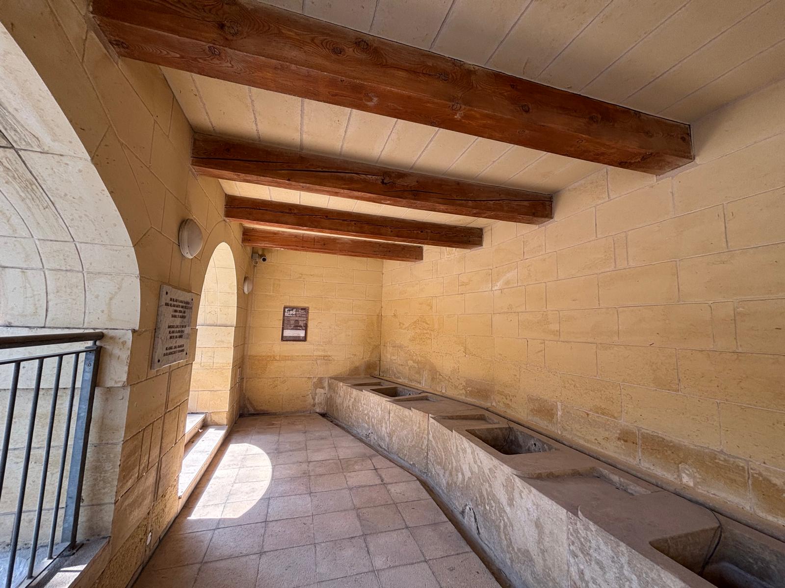 Interior view of Għajn tal-Ħasselin showing stone washing troughs aligned under a ceiling with heavy wooden beams