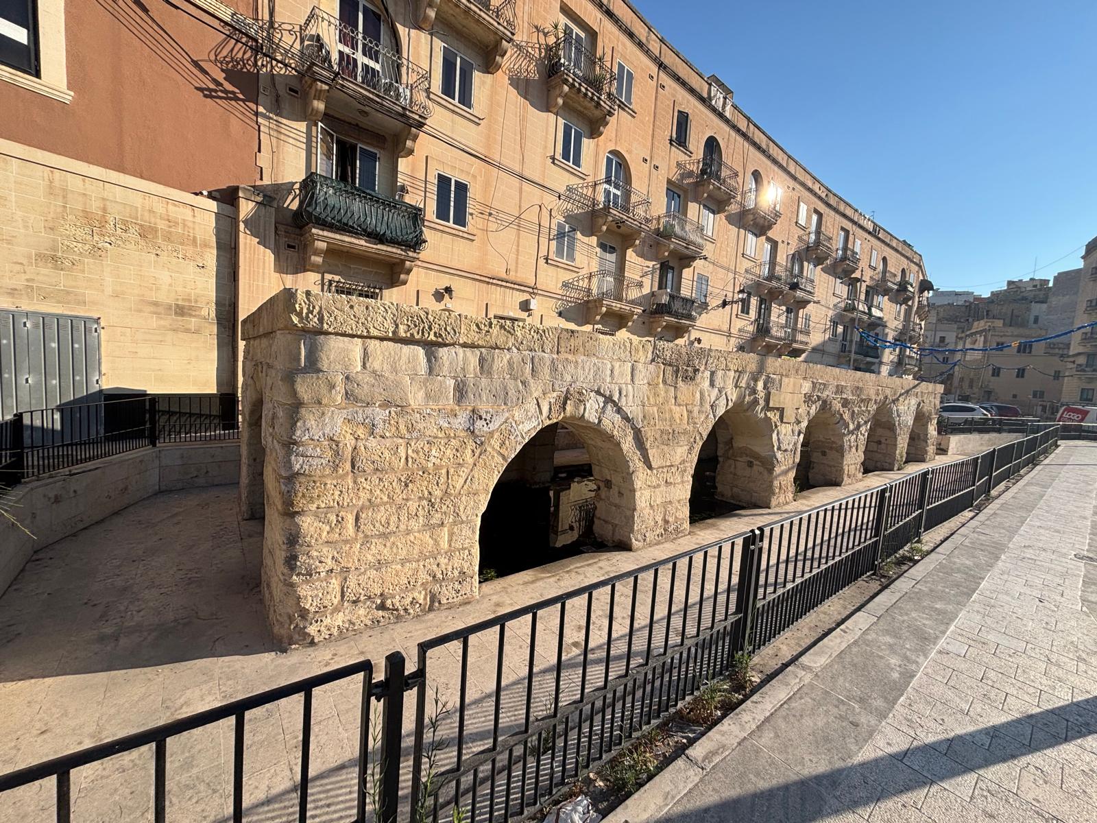 Wide angle road view of the complete arched Msida spring public wash house structure flanked by modern asphalt roads