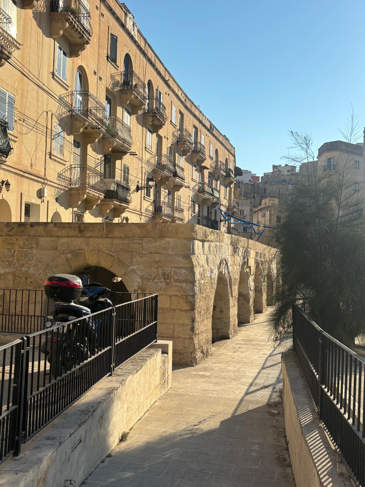 Pedestrian ramp and pavement pathway leading down to the entrance arches of the historic Msida spring