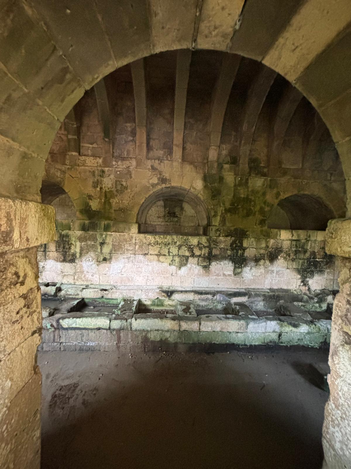 Interior view through a stone archway showing long stone washing troughs under large ribbed vaults