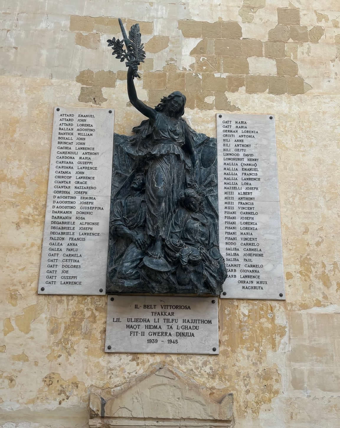 Overall view of the Birgu World War II memorial monument featuring bronze relief figures flanked by two large marble plaques listing casualties.